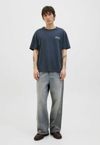 Mørkegrå bomulds T-shirt med korte ærmer og lille farverig grafik på brystet, parret med lysegrå vide denim jeans og brune sko.