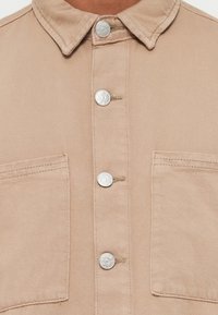 Beige denimskjorte med krave, sølvfarvede trykknapper og to forlommer. Har et struktureret stof med synlige søm.