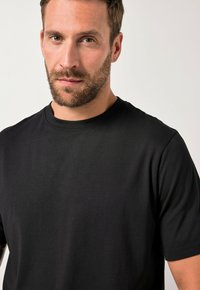 Hombre con barba y cabello corto castaño que lleva una camiseta negra de cuello redondo y mangas cortas, mirando hacia adelante contra un fondo claro.