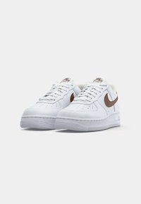 Par obu bijelih niskih tenisica s smeškim Nike swoosh logotipima i bijelim vezicama, prikazano iz prednjeg kuta na običnoj pozadini.