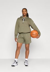 Haut à capuche olive avec cordon et poche avant, shorts assortis, baskets noir et blanc. Tenant un ballon de basket orange.