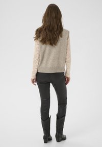 Frau mit langen braunen Haaren, die ein beiges Spitzenoberteil mit langen Ärmeln, einen grauen ärmellosen Strickpullover, schwarze Lederhosen und schwarze Stiefel trägt und von hinten zu sehen ist.