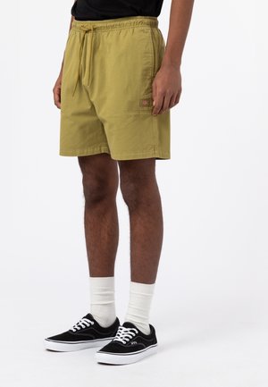 Persona vistiendo un pantalón corto con cordones amarillo mostaza, camiseta negra, calcetines blancos hasta la pantorrilla y zapatillas negras de baja corte, de pie sobre un fondo blanco.