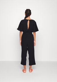 Mono negro con mangas cortas, cintura ceñida y detalle de ojal abierto en la espalda. Combinado con sandalias planas de color naranja brillante.