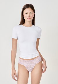T-shirt blanc ajusté à manches courtes associé à un string en dentelle rose présentant un motif floral et des bords festonnés, mettant en valeur la peau en dessous.