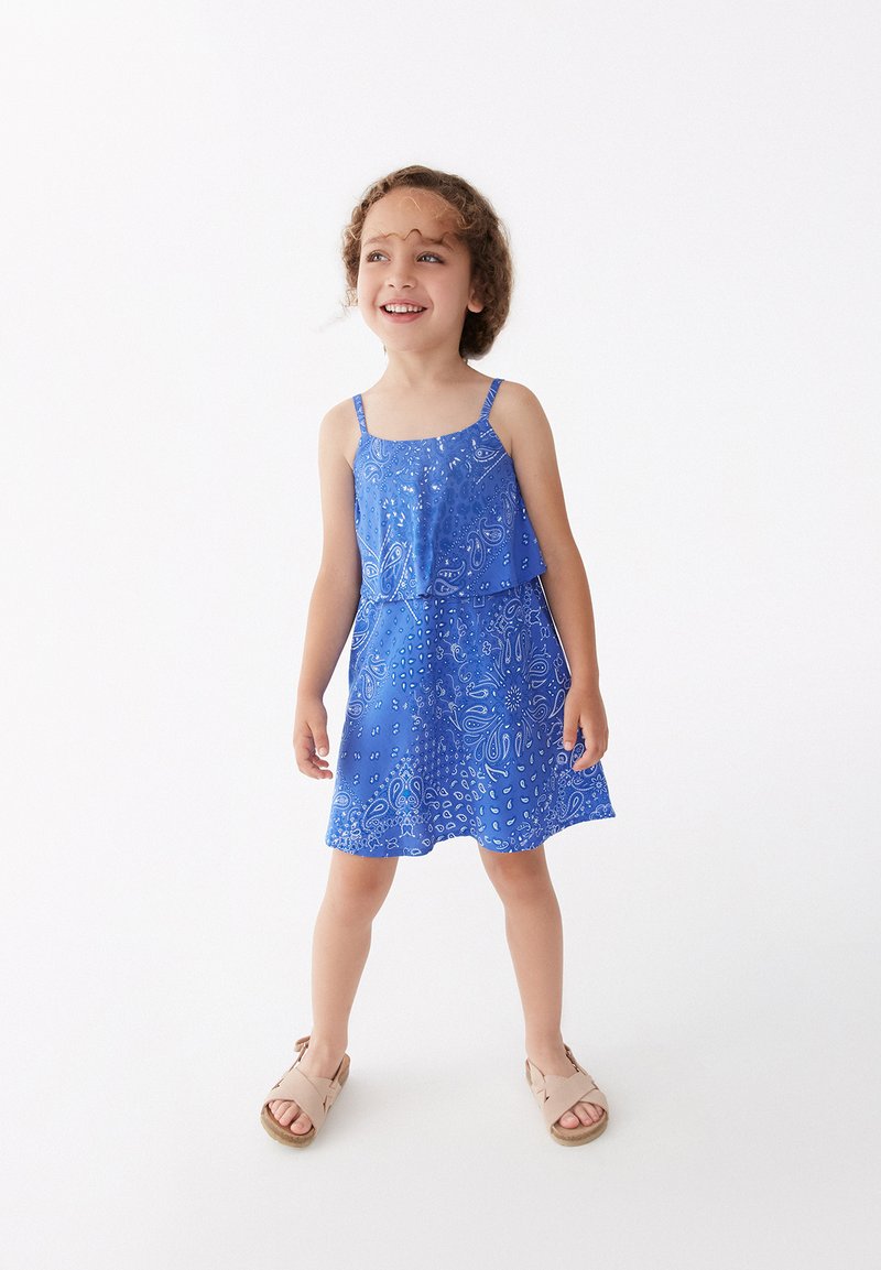 Vestido sin mangas azul con patrón de paisley, tela ligera, diseño en capas, largo hasta la rodilla, combinado con sandalias beige.