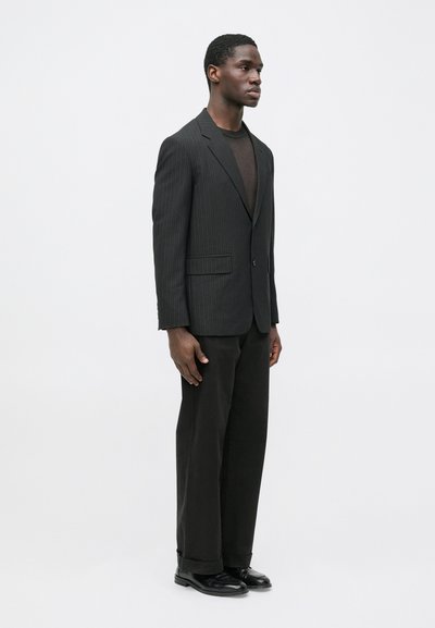 Čierny prúžkovaný oblekový sak s výrezovými klopami, doplnený tmavo hnedou košeľou a čiernymi nohavicami s vysokým pásom, dokončený čiernymi topánkami.