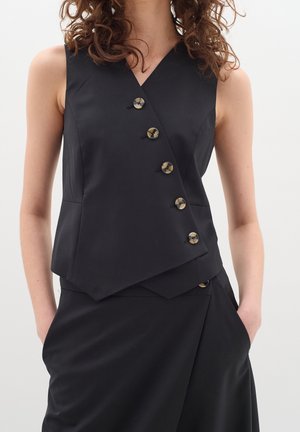 Veste noire sans manches asymétrique à cinq boutons, portée avec une jupe noire assortie, mains dans les poches de la jupe, cheveux bouclés visibles.