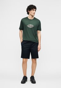 Νέος άνδρας με σγουρά μαλλιά που φοράει σκούρο πράσινο t-shirt με γραφικά, μαύρο σορτς και μαύρα αθλητικά παπούτσια, στέκεται με τα χέρια στις τσέπες.