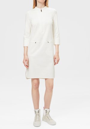 Kobieta w białej sukience za kolano z rękawami do połowy przedramienia, suwakiem z przodu, kieszeniami na zamek oraz białych wysokich sneakersach na sznurowadła z koronką.