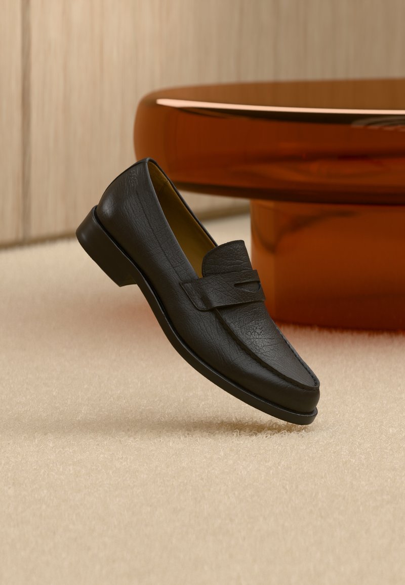 Mocassin en cuir noir flottant au-dessus d'un tapis beige avec une table ronde brune brillante en arrière-plan.