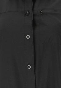 Zwart overhemd met aan de voorkant een knopenstrook met vier zwarte knopen en een borstzak met een klein ritssluitingtrekje aan de rechterkant.