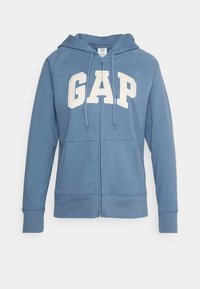 Felpa blu con zip realizzata in tessuto morbido, dotata di tasca a marsupio e grande logo bianco "GAP" sulla parte anteriore, con cappuccio regolabile tramite lacci.