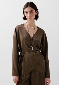 Top marrón con envoltura, confeccionado en tejido texturizado, con escote en V, mangas largas y cintura anudada con elementos decorativos y un colgante plateado.