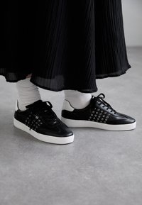 Sneakers neri con accenti di occhielli rotondi, una parte superiore in pelle liscia e una suola in gomma bianca. Abbinati a calzini bianchi e una gonna plissettata nera.