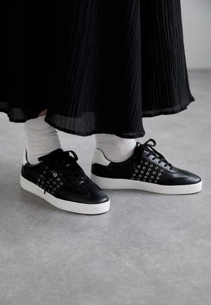 Black sneakers with round eyelet accents, a smooth leather upper, and a white rubber sole. Paired with white socks and a black pleated skirt.