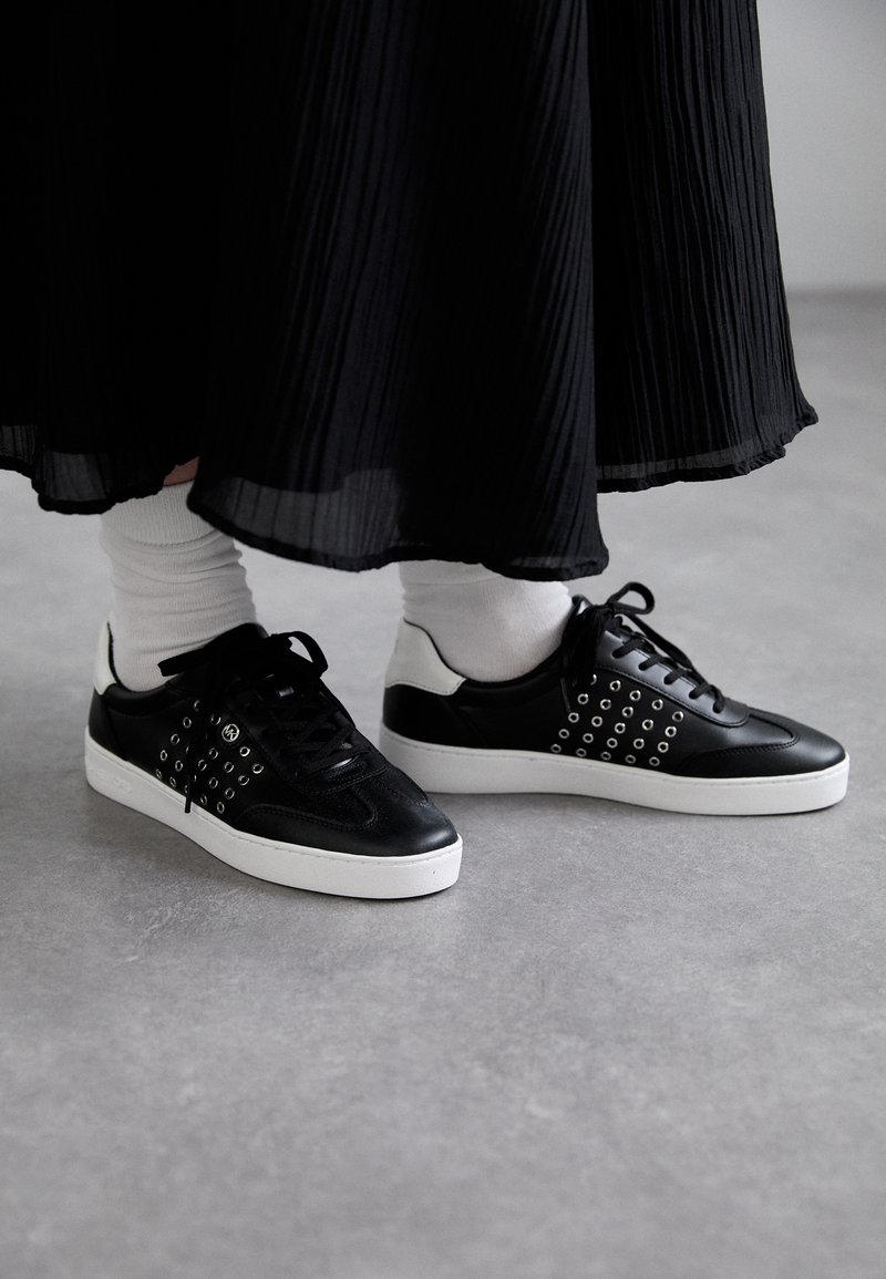 Sneakers neri con accenti di occhielli rotondi, una parte superiore in pelle liscia e una suola in gomma bianca. Abbinati a calzini bianchi e una gonna plissettata nera.