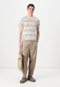 Striped t-shirt in off-white with green, blue, and orange lines; beige relaxed-fit trousers; gray footwear; holding a beige bag.