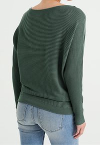 Grön rippad tröja med avslappnad passform, bred halsringning och långa ärmar, kombinerad med ljusblå jeans. Tyget har en strukturerad yta.