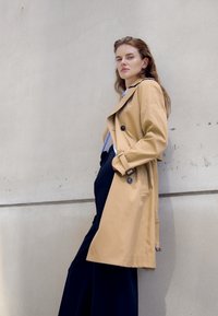 Hellebeige Trenchcoat mit einem Gürtel in der Taille, Knopfakzenten und einem breiten Kragen, kombiniert mit dunklen, weiten Hosen vor einer hellen Wand.