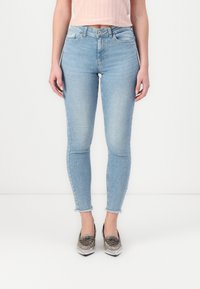 Világoskék skinny farmernadrág, melynek szélén rojtos hem van, öt zsebbel és cipzáros, gombbal záródó fazonnal. Fémes loaferekkel viselve.