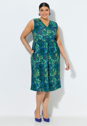 Smilende kvinde iført ærmeløs, knælængde blå kjole med grønt bladprint, blå øreringe og blå høje hæle, stående mod en hvid væg.