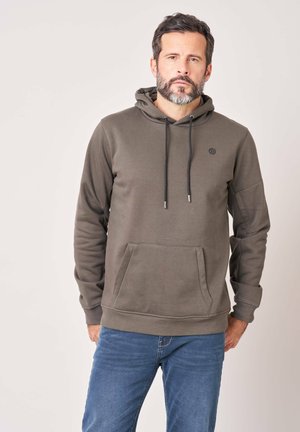 Homme avec une barbe portant un sweat à capuche vert olive et un jean bleu, debout devant un fond uni.