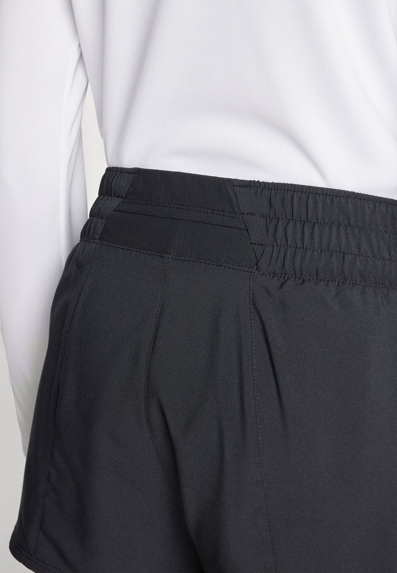 Zwarte sportshorts met een elastische tailleband en horizontale stiksels, met een gladde textuur, gedeeltelijk zichtbaar naast een witte longsleeve.