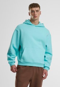 Tirkizna oversized dukserica s kapuljačom, sa vezicom, kenguruh džepom, rebrastim manžetama, izrađena od mekog materijala. Uparena s smešnim hlačama.