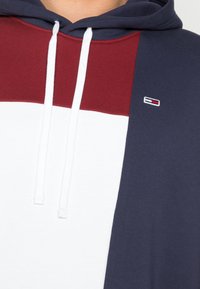Kapuzensweatshirt mit einem Farbblockdesign, das Abschnitte in Marineblau, Weiß und Burgunderrot aufweist. Weicher Stoff mit weißen Kordeln und einem Logo.