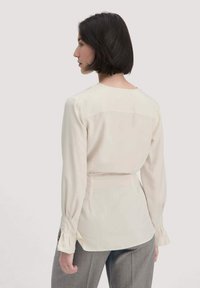 Donna con capelli corti scuri che indossa una camicia a maniche lunghe color crema e pantaloni grigi, rivolta di lato su uno sfondo chiaro.