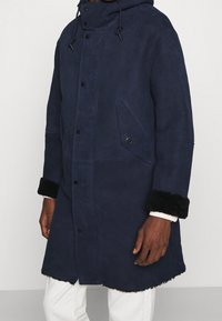Manteau en suède bleu marine avec capuche, munis de poignets en fausse fourrure noire, fermeture à boutons et poches avant. Texture douce avec une coupe mi-longue.