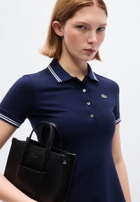 Jeune femme en polo bleu marine à rayures blanches tenant un sac à main texturé noir devant un fond uni.