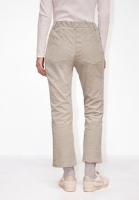 Personne portant un pantalon beige en velours côtelé, des chaussettes gris clair, des baskets beiges et une chemise à manches longues rose clair, debout de dos sur un fond blanc.