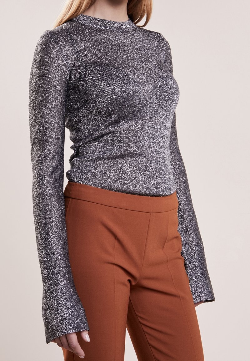 Femme portant un haut argenté pailleté ajusté à manches longues avec un pantalon marron taille haute sur un fond uni.