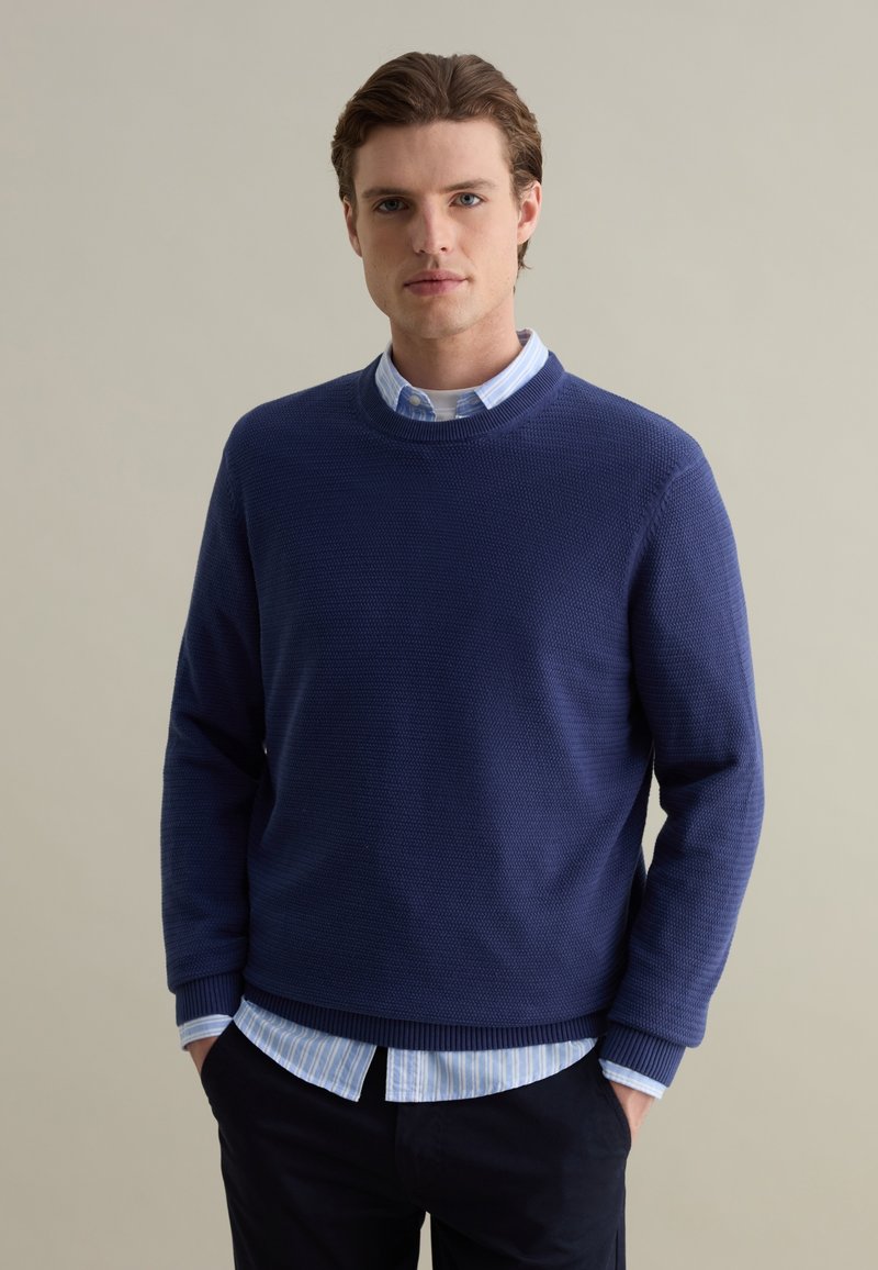 Navy blue knit sweater with a textured design, round neck, and ribbed cuffs, layered over a light blue striped collared shirt.