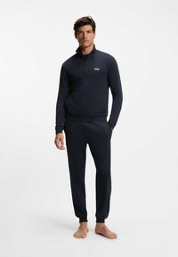 Tummansininen vetoketjullinen takki korkealla kauluksella, yhdistetty samansävyisiin housuihin. Pehmeä kangas, istuva malli ja logodetalji rinnassa.