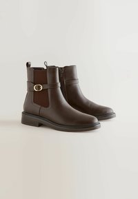 Bottes chelsea en cuir marron avec une fermeture éclair latérale, un panneau texturé et un détail de sangle avec accent doré. Talon légèrement rehaussé et finition lisse.