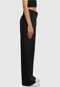 Personne portant un pantalon noir taille haute à jambes larges et des baskets blanches, debout de profil, avec la main détendue le long du corps.