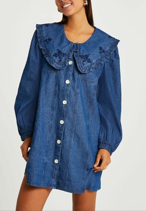 Femme portant une robe chemise en denim bleu à boutons, avec de longues manches bouffantes et un grand col à volants orné de broderies florales foncées.