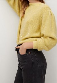 Gelber Strickpullover mit strukturiertem Finish und dezenten Pailletten, kombiniert mit schwarzen Jeans. Der Pullover hat einen runden Halsausschnitt und puffige Ärmel.