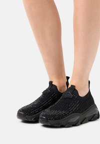 Black slip-on sneakers with a mesh upper featuring a textured pattern and chunky black sole. Includes logo accent on the tongue.