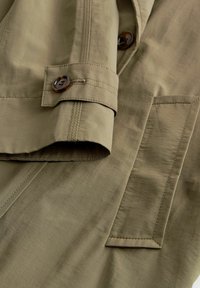 Licht olijfgroene jasstof met een gladde textuur, voorzien van een knoopmanchet, zichtbare stiksels en een patchzakdetail.