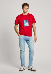 Red cotton t-shirt featuring a blue graphic print labeled "Pepe Jeans." Worn with light blue jeans and white sneakers with black accents.