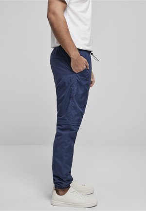Homme portant un pantalon de jogging bleu marine avec des poignets élastiques et des baskets blanches, debout avec une main dans la poche sur un fond uni.
