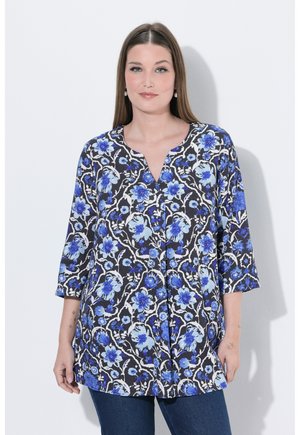 Kobieta o prostych brązowych włosach, ubrana w niebieską, kwiatową bluzkę z rękawami do łokcia i ciemne dżinsy, stojąca na białym tle.