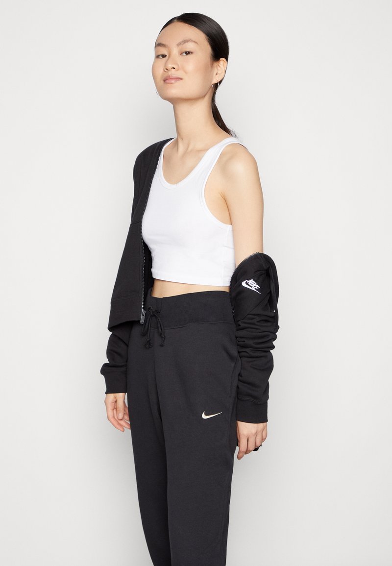 Joggers negros de Nike con cintura elástica y logo, combinados con una camiseta de tirantes corta blanca y una chaqueta de sudadera negra con cremallera, usada parcialmente.
