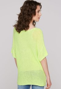 Neon yellow oversized knit top with short sleeves, open weave texture, and a rounded neckline, paired with light blue jeans.