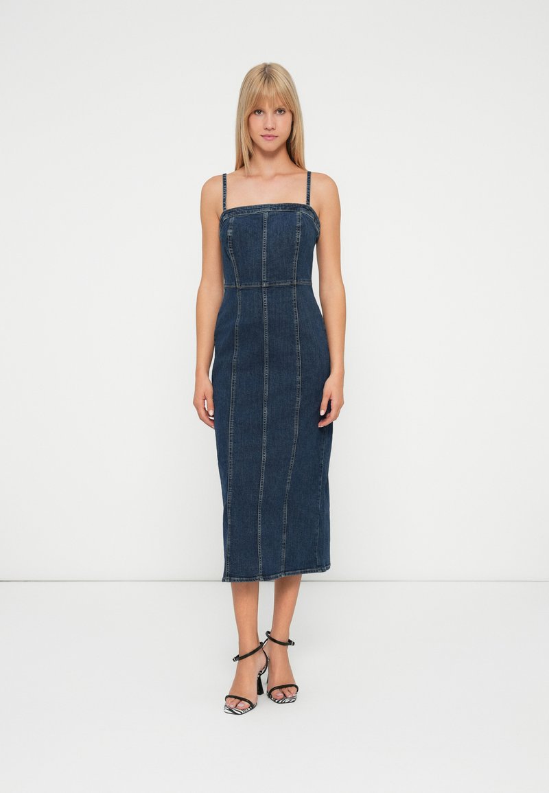 Vrouw staat rechtop met het gezicht naar voren, gekleed in een aansluitende donkerblauwe denim midi-jurk met dunne bandjes en zwarte hoge hakken in een witte studio.