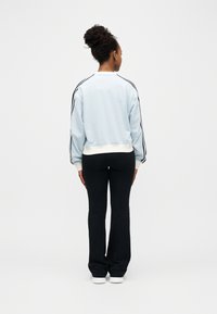 Femme aux cheveux bouclés attachés en chignon, portant un sweat-shirt bleu clair avec des rayures noires sur les manches, un pantalon noir et des baskets blanches, debout de dos.